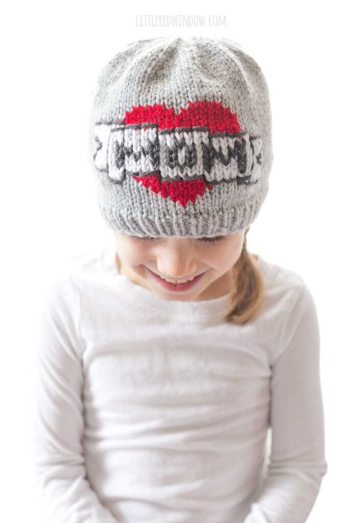 girl in a white shirt in front of a white background looking down at her lap and wearing a gray knit beanie with the classic mom tattoo motif on the front a red heart with a white banner in front that reads MOM