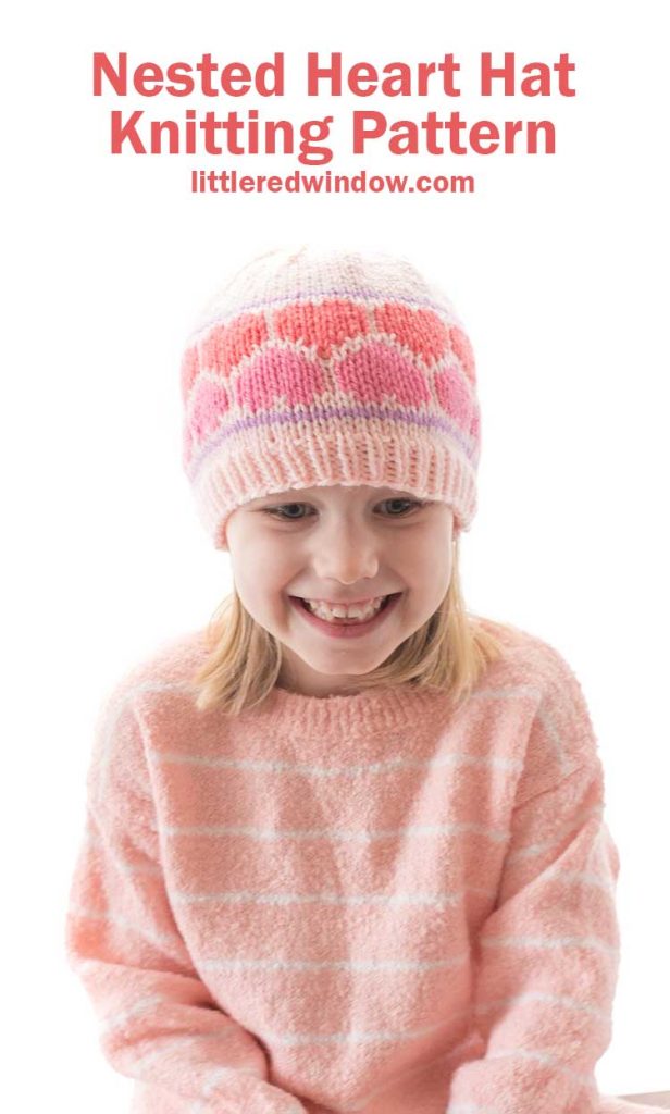 smiling girl in pink sweater wearing a pink knit hat with multicolored pink knit hearts around the middle