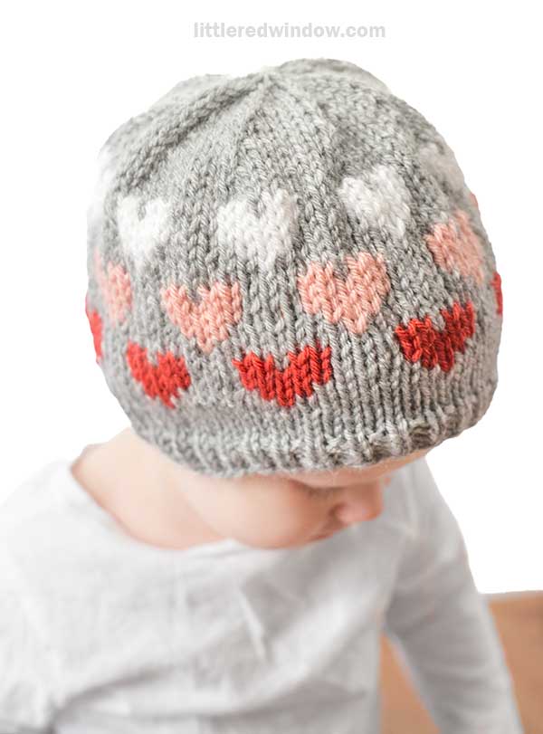 A child wearing a gray heart hat is lost in thought, gazing downward. The knit masterpiece features a delightful pattern of red, pink, and white hearts, capturing the essence of craftsmanship and style.