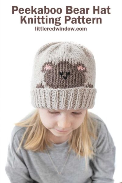 blonde girl in gray shirt looking down wearing a tan knit hat with a folded brim that has a brown teddy bear head peeking over the brim