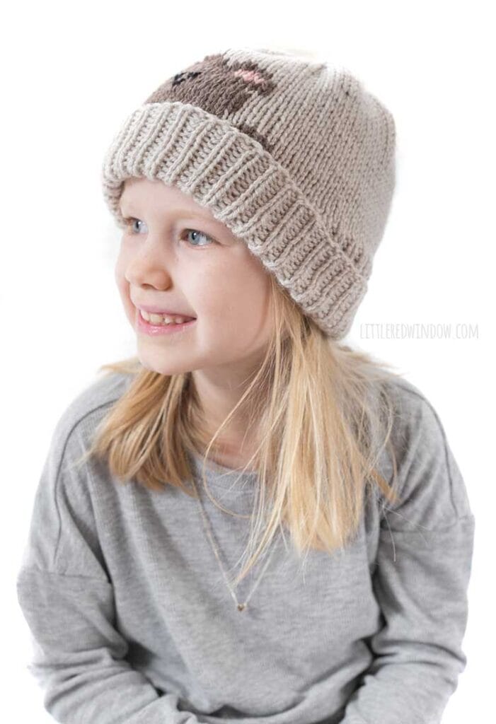 smiling blonde girl in gray shirt looking to the left wearing a tan knit hat with a folded brim that has a brown teddy bear head peeking over the brim