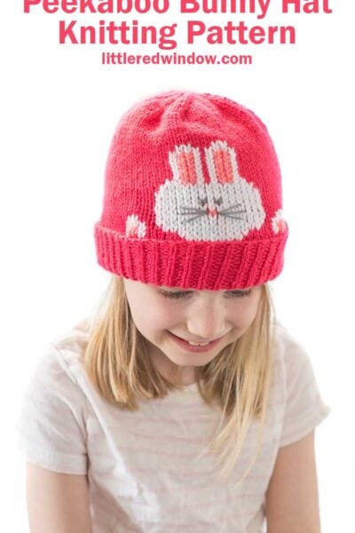 smiling girl in striped short sleeved shirt wearing a hot pink knit hat that has a white bunny face and paws peeking out over the folded brim looking down at her lap
