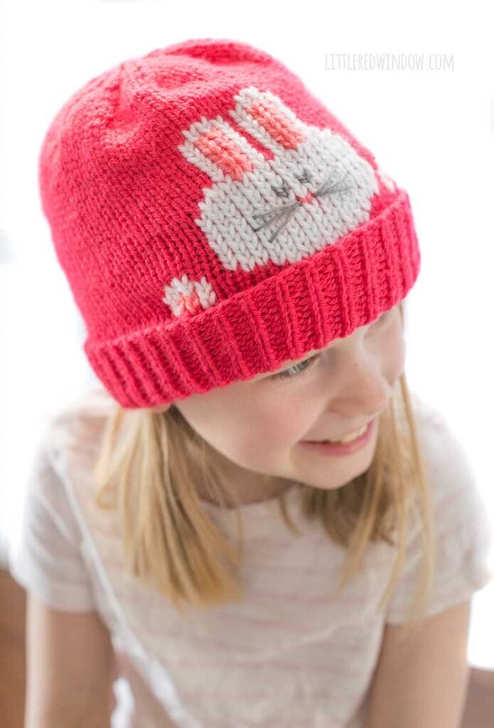 view from slightly above smiling girl in striped short sleeved shirt wearing a hot pink knit hat that has a white bunny face and paws peeking out over the folded brim looking off to the right