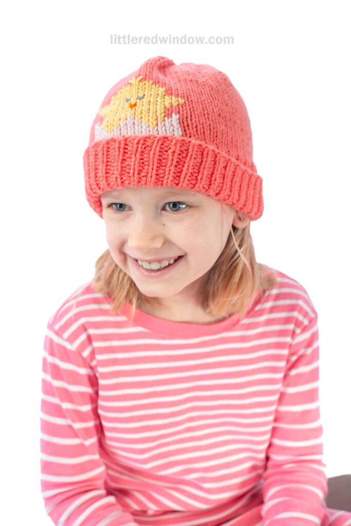 A smiling girl wearing a knitted coral beanie adorned with a yellow spring chick design, a chick hat knitting pattern, complements her coral and white striped shirt. She gazes to her right against a plain white background.