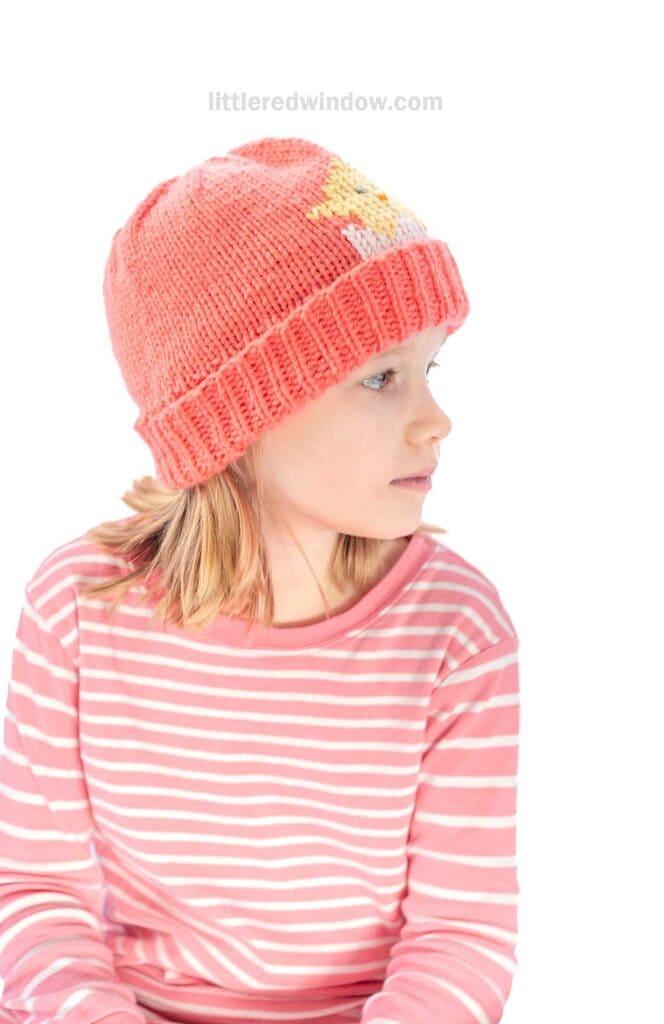 A child wearing a coral knit hat with a chick knitting pattern, with a yellow and white design, looks to the side. Their shoulder-length hair complements a matching coral and white striped shirt against a plain white background.