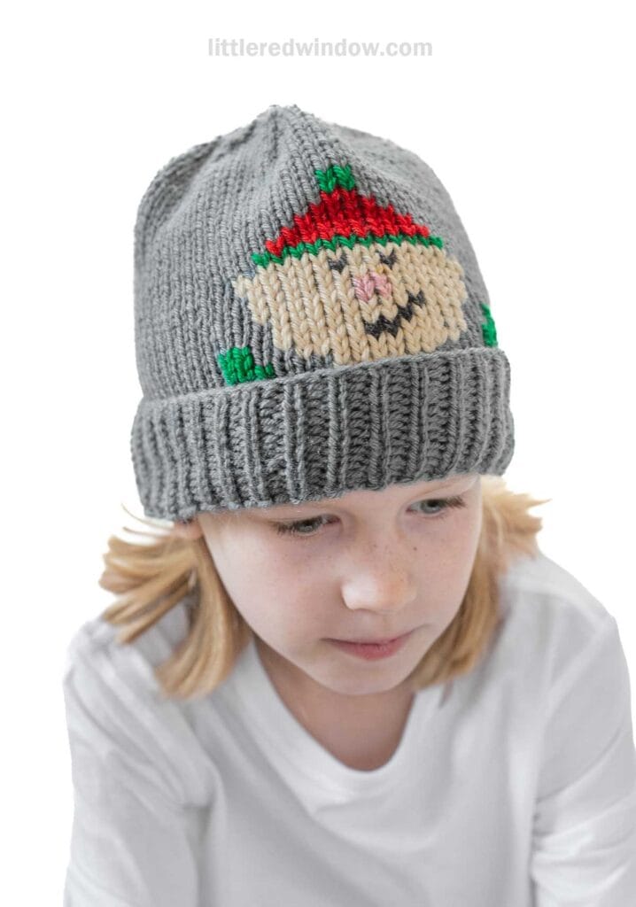 A child wearing a gray knit hat featuring a Santa design, reminiscent of an elf hat knitting pattern. The child is slightly looking down, dressed in a white shirt against a plain white background.