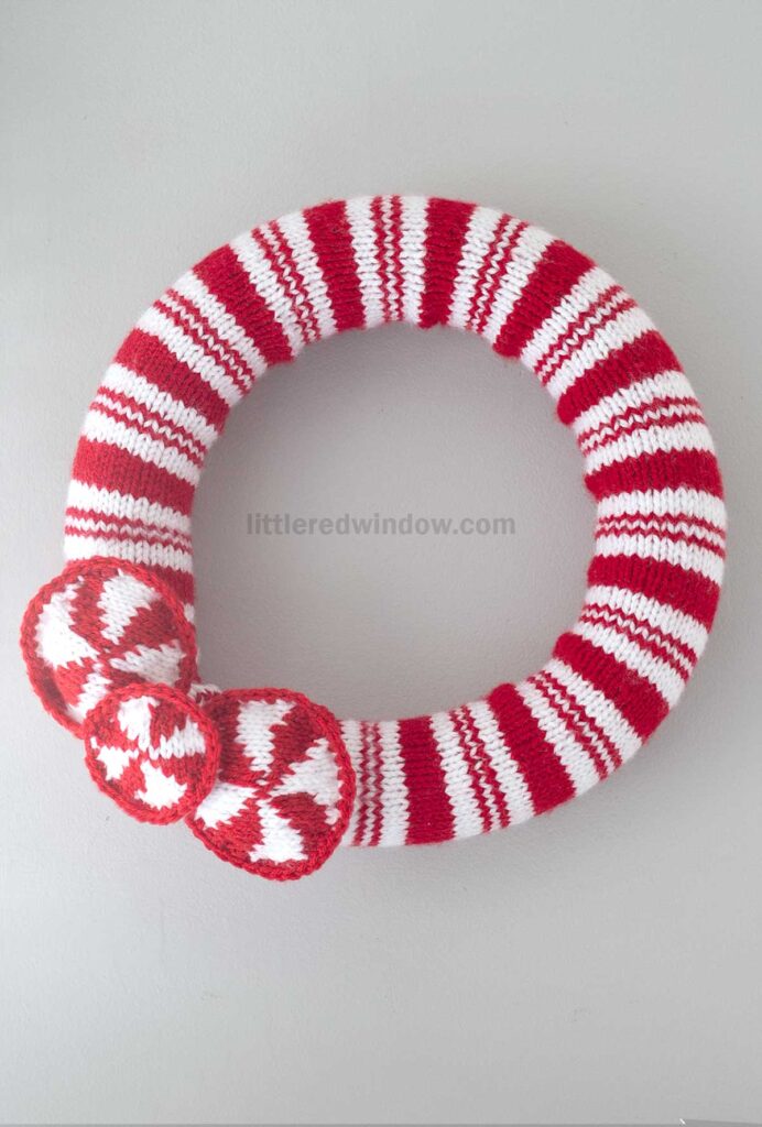 A round holly wreath wrapped in red and white striped yarn with three knitted red and white peppermint candy decorations attached, showcasing a charming knitting pattern, hangs elegantly on a light gray wall.