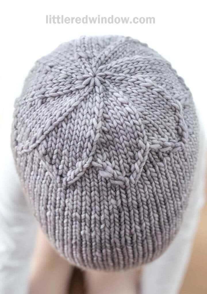 The image shows a close-up top view of a person's head wearing a Petal Hat with intricate star-shaped stitching. The hat is light gray in color, and the background is blurred, focusing attention on the detailed knit pattern.