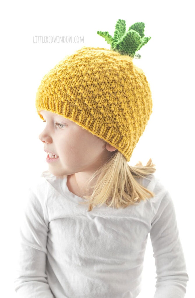 child in white shirt wearing a knit pineapple hat looking off to the left in front of a white background