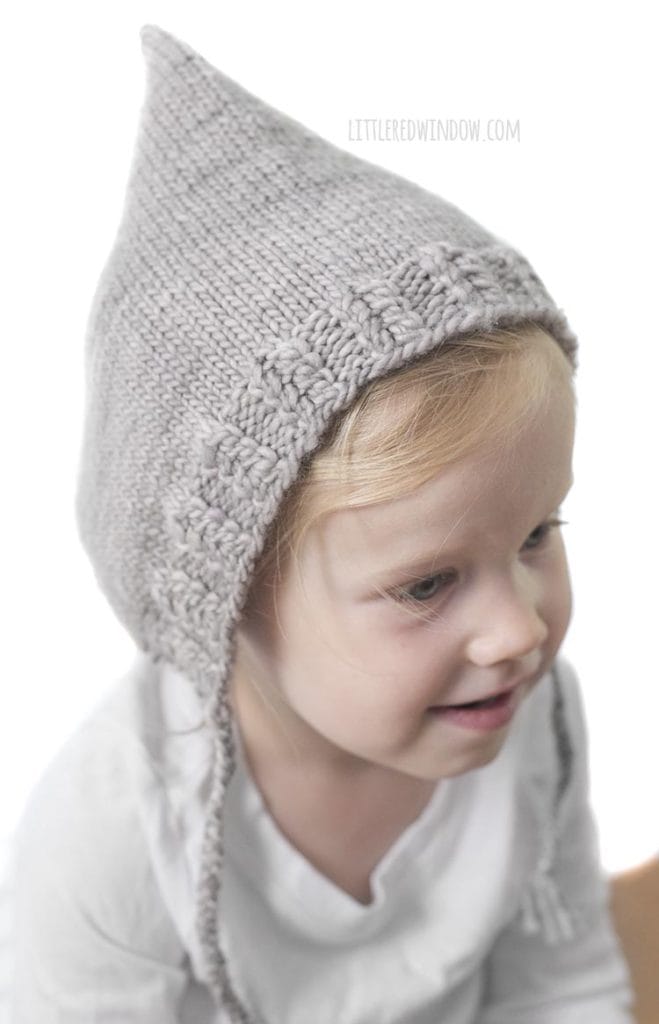 little girl in white shirt wearing tan knit pixie bonnet with braided chin ties looking off to the right