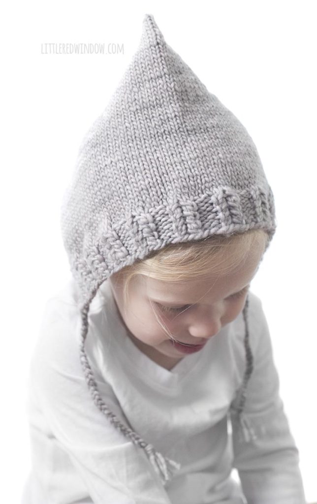 little girl in white shirt wearing tan knit pixie bonnet with braided chin ties looking down and to the right