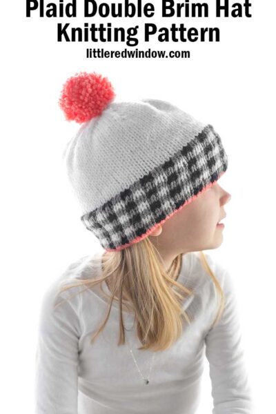girl in white shirt wearing a white knit hat the black and white plaid brim and pink pom pom on top looking off to the right