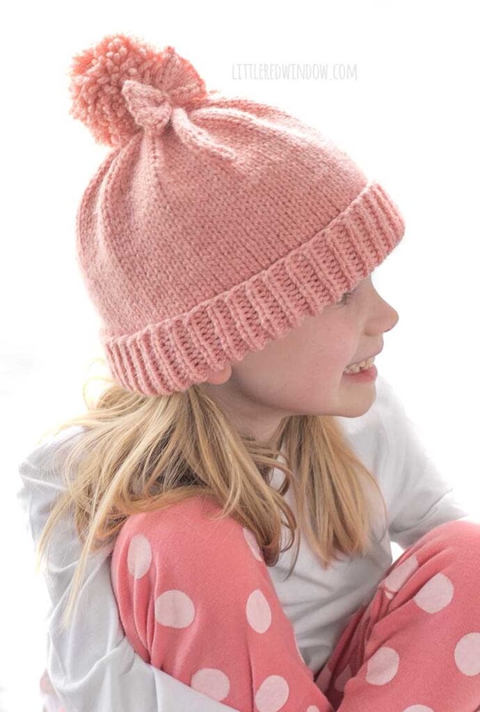 girl in white shirt and pink pants sitting cross legged looking off to the right and wearing a medium pink knit hat with a folded brim a big pom pom on top and a bow tied around the pom