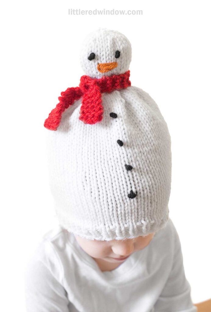 A child sports a charming snowman hat, knitted in white with black buttons and a cheerful red scarf. The top mirrors a snowman's head, complete with embroidered eyes and a friendly smile.