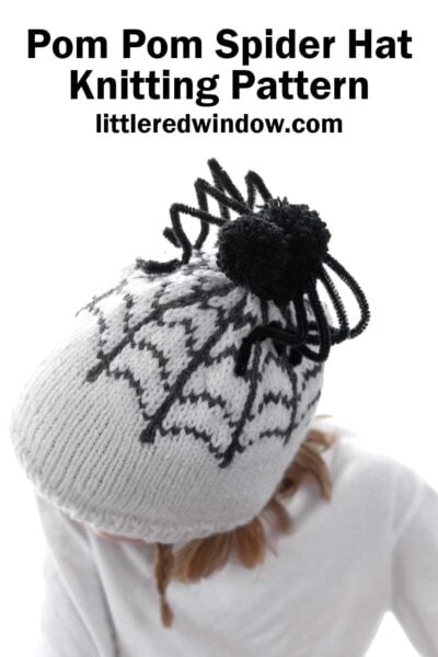 A child wearing a white knitted hat featuring a black spider web design and a black pom pom on top. The text "Pom Pom Spider Hat Knitting Pattern" and "littleredwindow.com" appears at the top of the image. The child is looking down, and the background is plain white.