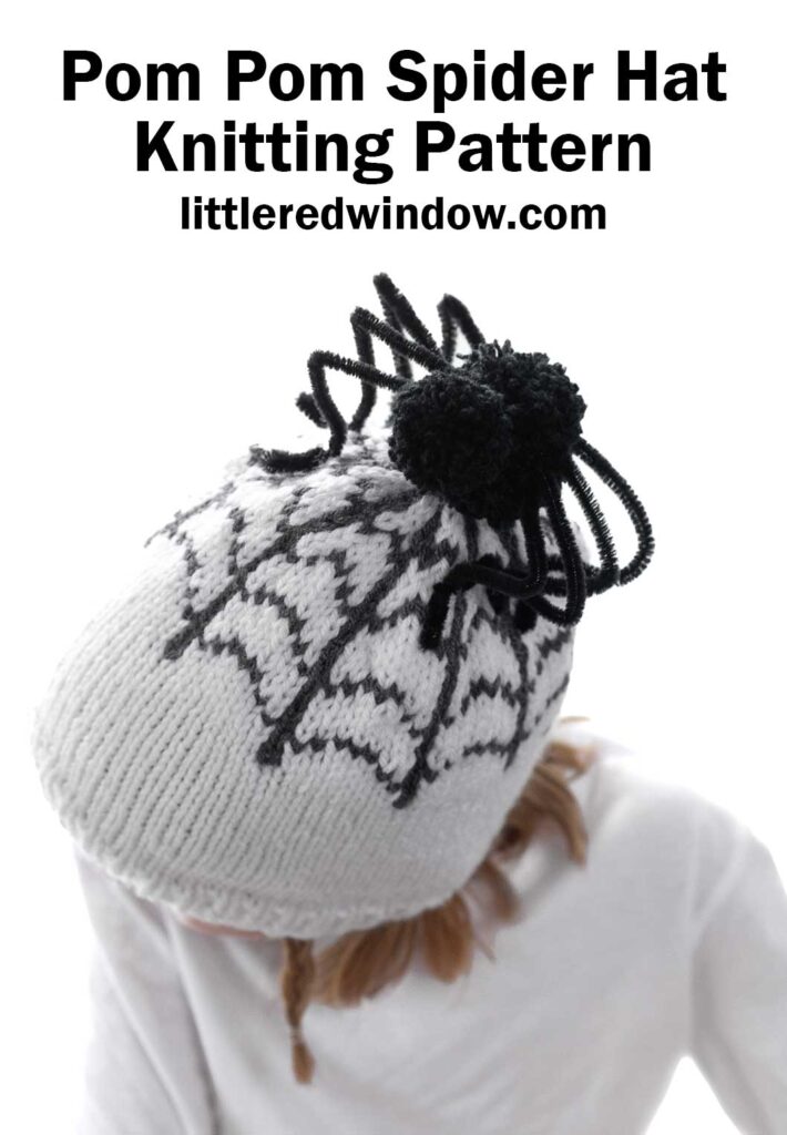 A child wearing a white knitted hat featuring a black spider web design and a black pom pom on top. The text