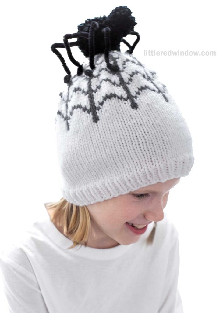A child with blonde hair is wearing a white knitted hat featuring a large black spider design and a pom-pom on top. The child is smiling and dressed in a white shirt.