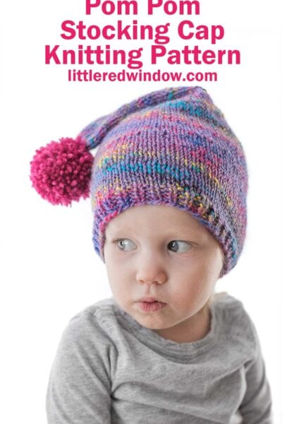 child in gray shirt with pursed lips wearing a purple speckled knit stocking cap with magenta pom pom hanging to the side while they look to the left