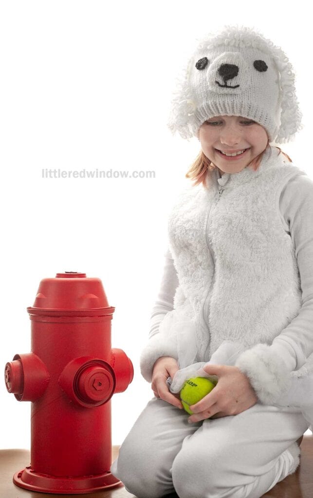 A child in a fluffy white outfit resembling a poodle, wearing a matching knit hat with poodle ears, smiles while holding a green tennis ball. A red fire hydrant is beside the child.