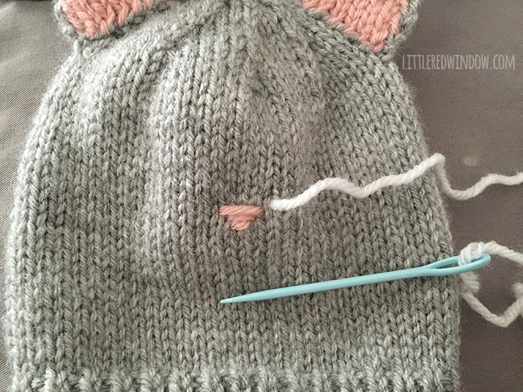 Cat Hat With white yarn on a blue yarn needle ready to stitch whiskers on the cat hat