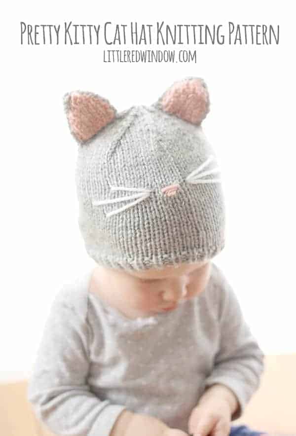 Baby in gray onesie wearing a gray knit cat hat with pink inner ears and a pink nose and whiskers looking down at their hands