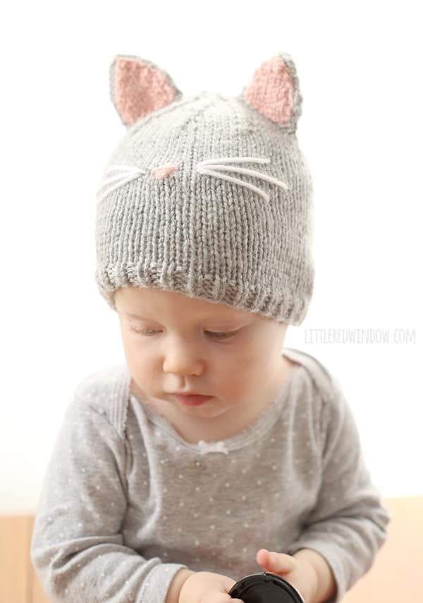 Baby in gray onsie holding a camera lens cover and wearing a gray knit cat hat with gray and pink cat ears and whiskers on the front looking down and to the left