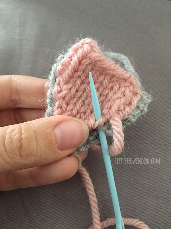 Hand holding the knit parts of the cat ear with a blue plastic yarn needle sewing the parts together in front of a gray background
