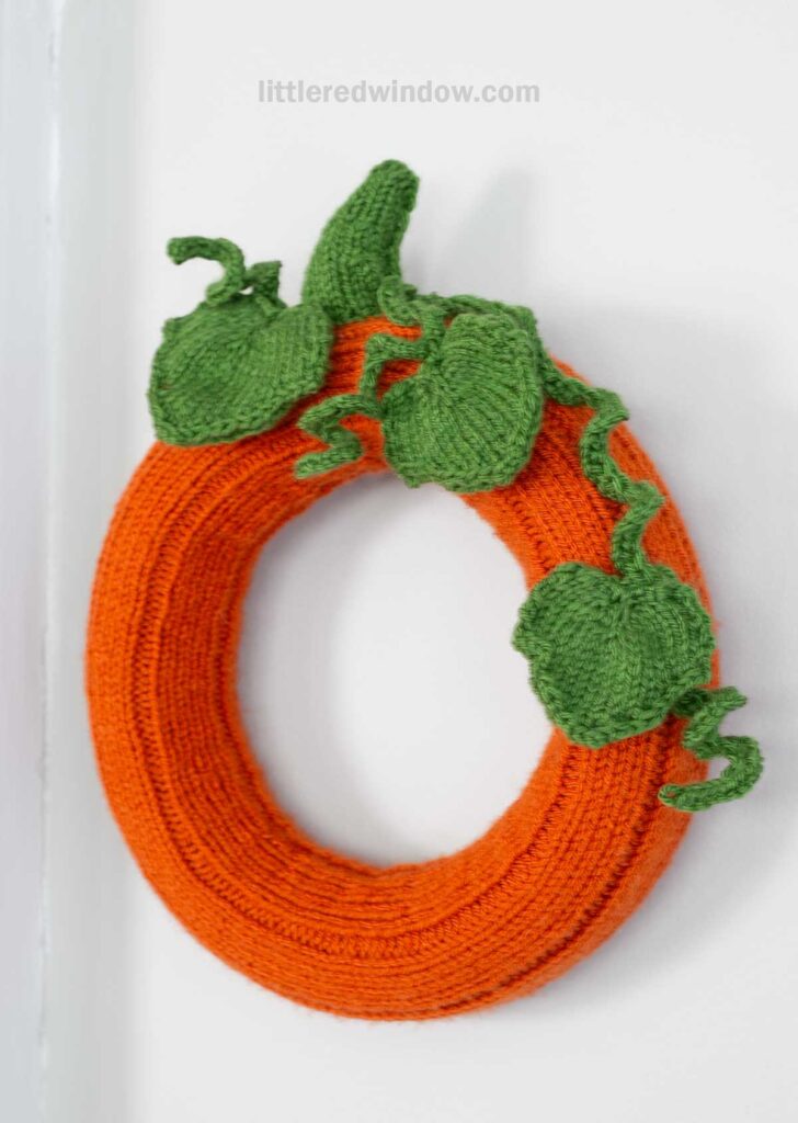 A knitted pumpkin wreath wrapped in bright orange yarn with green leaves and vines, resembling a pumpkin, hangs on a white wall.