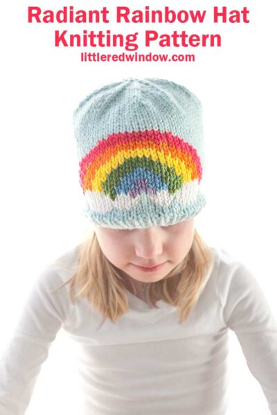 A child dons a charming light blue knitted hat adorned with a vibrant rainbow pattern. Above, the text declares: "Radiant Rainbow Hat Knitting Pattern - littleredwindow.com." The cheerful ensemble is complemented by the child's light-colored shirt.