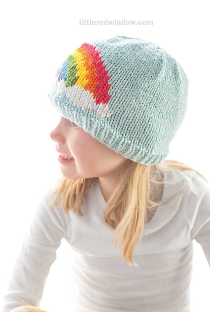 A child sported a delightful rainbow hat, crafted from a charming knitting pattern. The light blue knit hat featured a vibrant rainbow on the side as the child faced sideways, dressed in a white long-sleeve shirt against a bright and softly blurred background.