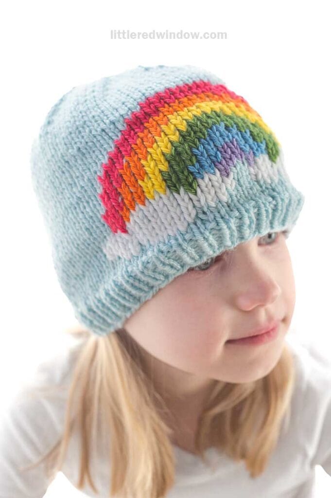 A child wearing a charming knitted hat adorned with a vibrant rainbow pattern gazes off to the side, dressed in a crisp white shirt. The background is a gentle, soft white.