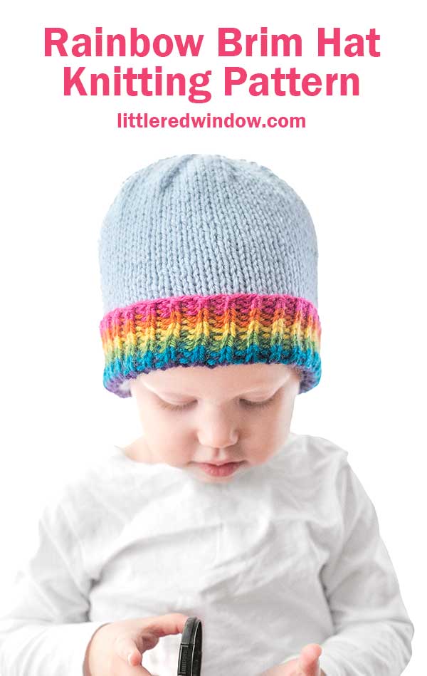 A baby in a charming knitted hat featuring a light blue top and a vibrant rainbow brim gazes down, clutching an object. Capturing the essence of this delightful