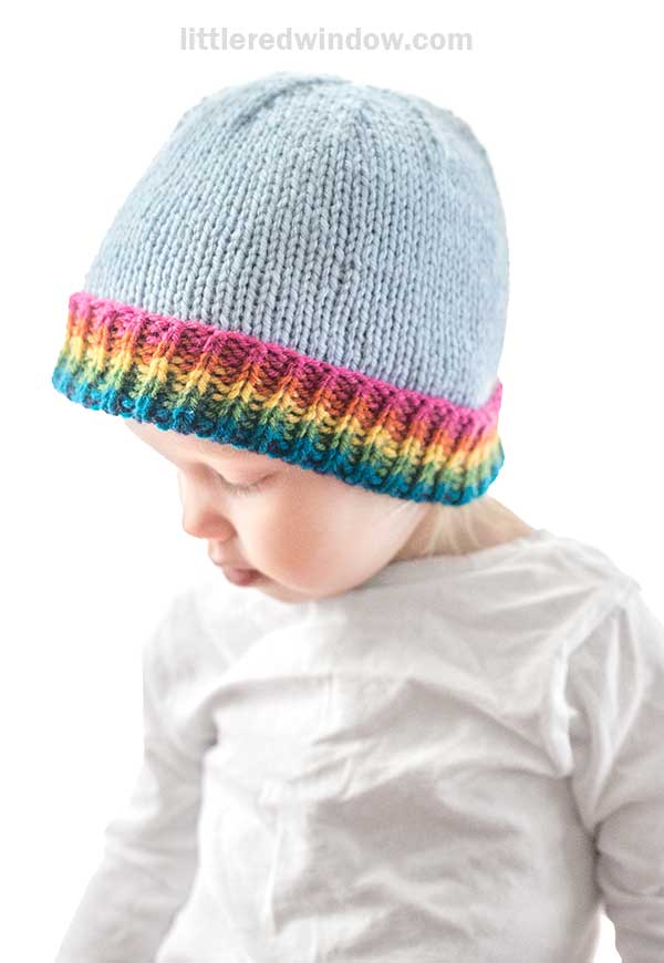 A baby wearing a light blue knitted hat with a vibrant rainbow brim gazes downwards. The little one is dressed in a crisp white shirt, set against a plain white background.