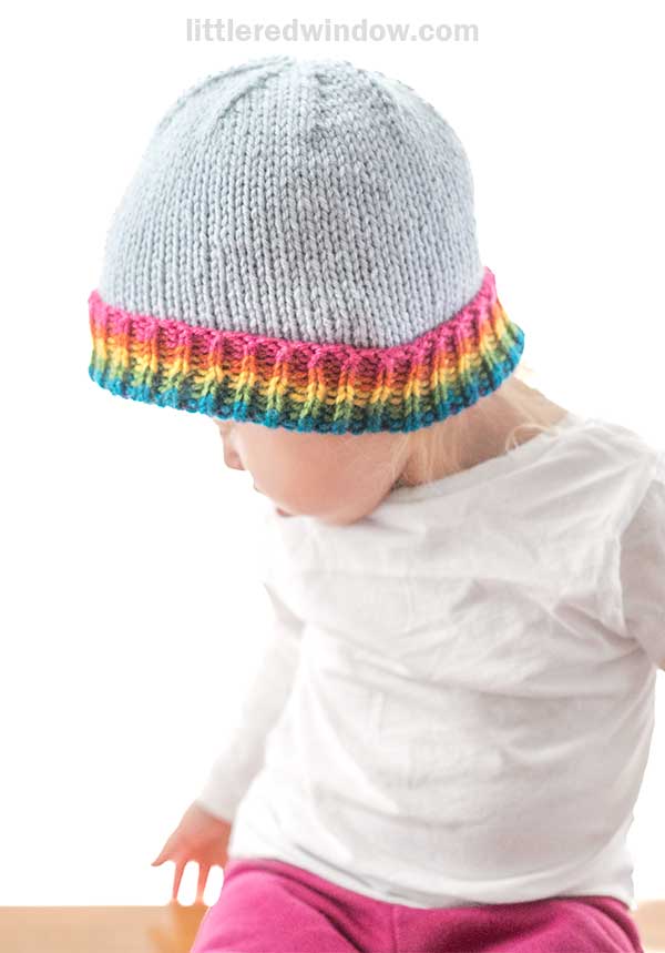 A child is wearing a light gray hat with a vibrant rainbow brim. Dressed in a white shirt and pink pants, the child gazes downward against a plain white background.