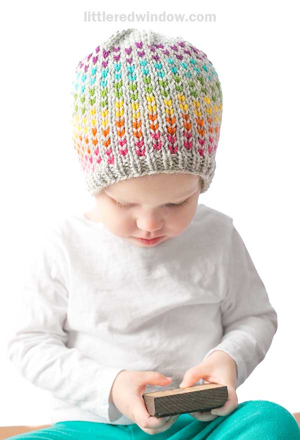 A child in a white shirt is wearing a colorful, knit hat with a patterned design featuring rainbow dots. They are looking down and holding a rectangular object against the plain white background.