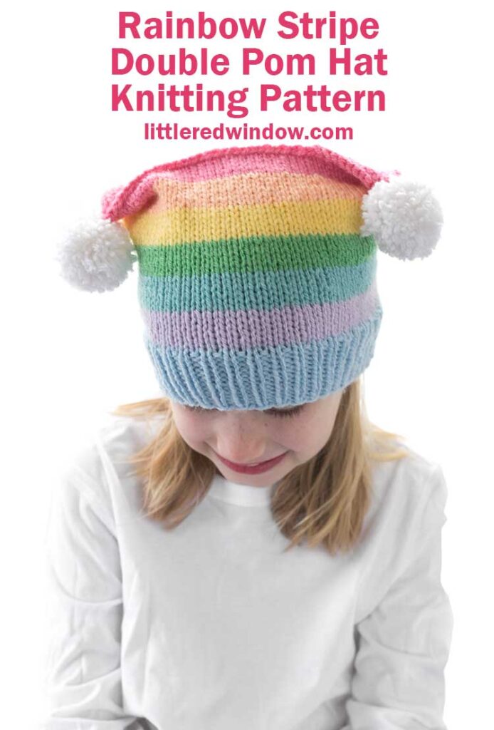 girl in white shirt wearing a rainbow striped hat with two white pom poms on the top corners looking down at her lap