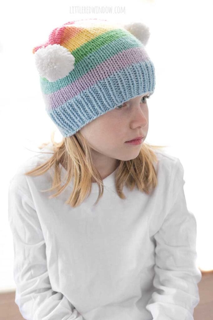 girl in white shirt wearing a rainbow striped hat with two white pom poms on the top corners looking off to the right