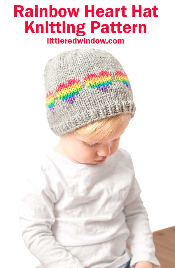 A toddler in a gray Rainbow Hearts Hat, adorned with vibrant heart patterns, looks down thoughtfully. Above them, the text reads 