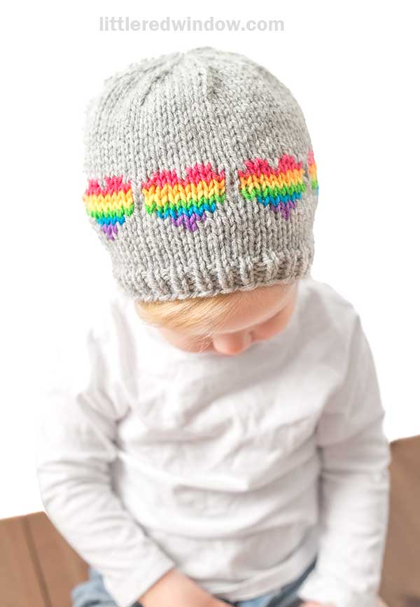 A child with a Rainbow Hearts Hat, knitted in gray and adorned with vivid colors, gazes downward in a simple white shirt against a plain backdrop.