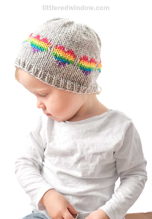 A child wearing a whimsical rainbow hearts hat sits with light hair cascading over a white long-sleeve shirt. Their gaze drifts down and slightly to the side, contrasting softly against the plain white background.