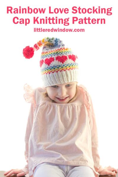 A child in a colorful stocking cap adorned with red hearts beams with joy. Above, the text reads, "Rainbow Love Stocking Cap Knitting Pattern," and "littleredwindow.com." The child is dressed in a light pink long-sleeve shirt.