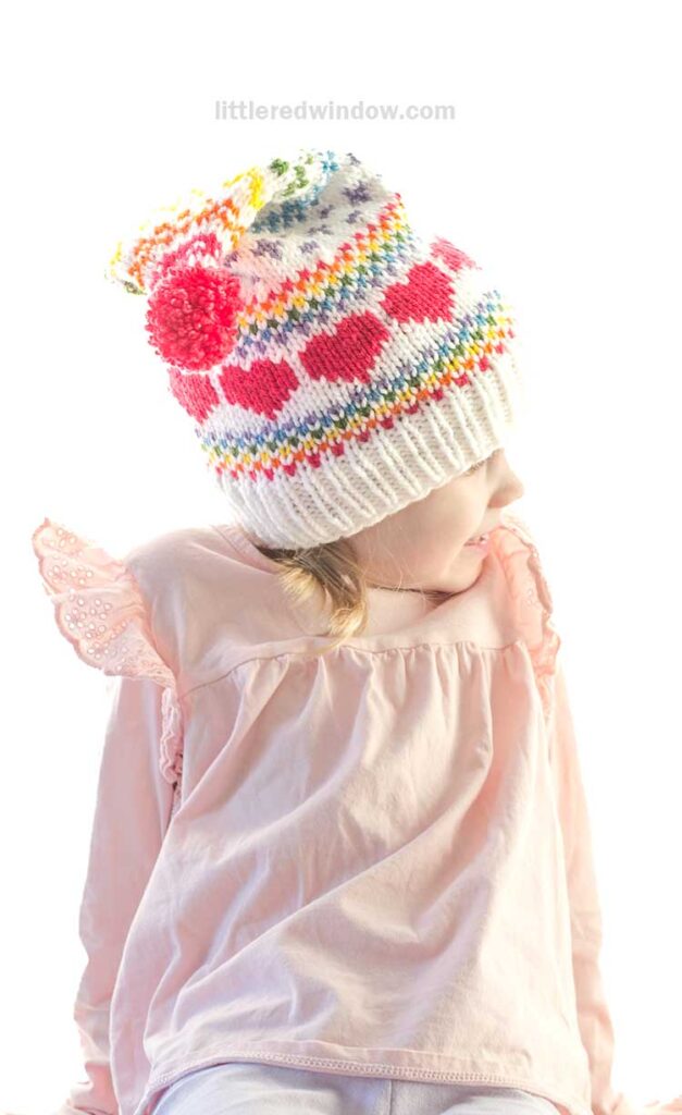 Child in a light pink top with lace sleeve details, wearing a colorful stocking cap featuring a knitting pattern of red hearts, stripes, and a pom-pom on top, looking to the side.