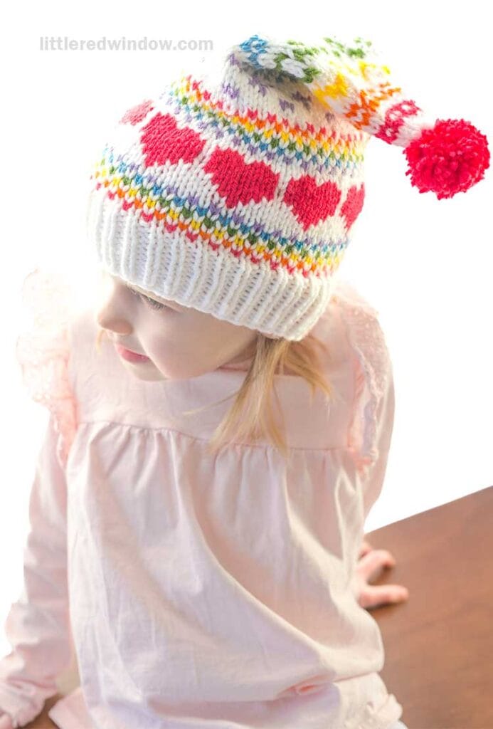 A child sits on a wooden surface, sporting a delightful white stocking cap adorned with colorful hearts and a playful pom-pom. The knitting pattern features red, pink, and rainbow designs. Complementing the hat, the child is dressed in a light pink top with charming ruffled sleeves.