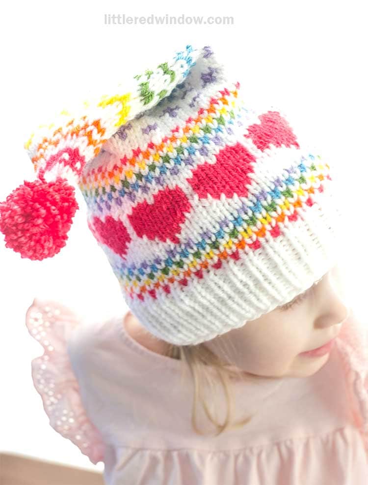 A child wearing a colorful stocking cap with red heart patterns, rainbow stripes, and a multicolored pom-pom on top delights in their look. The charming knit hat pairs perfectly with the child's pale pink shirt adorned with ruffled sleeves.