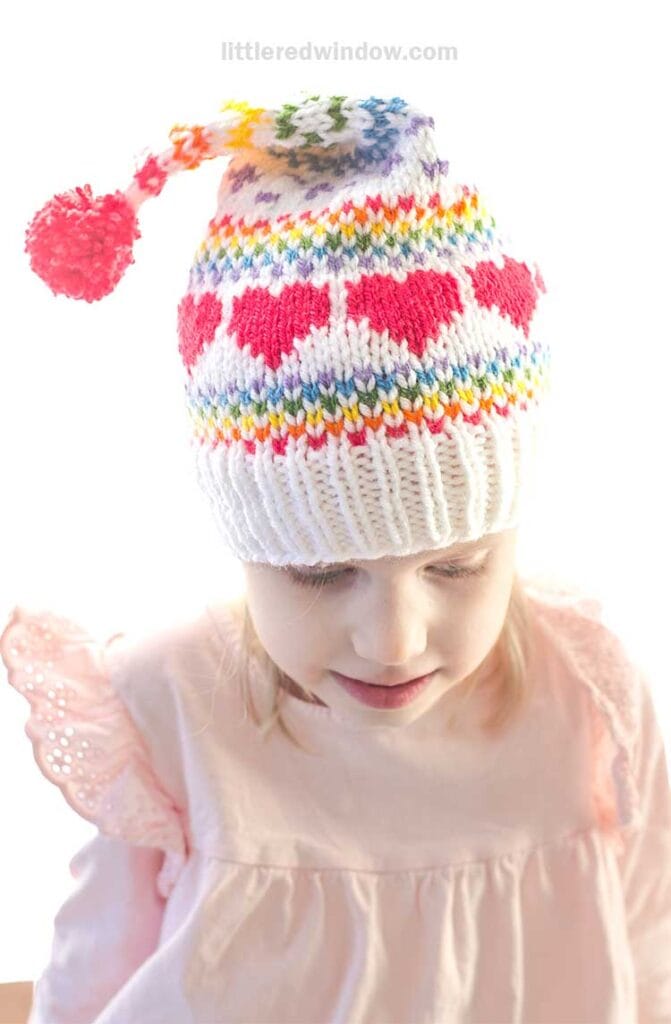 A child wearing a colorful stocking cap knitting pattern with red heart designs and a vibrant pom-pom on top. The hat, adorned with rainbow stripes, perfectly complements the child’s pink blouse as they gaze downward.