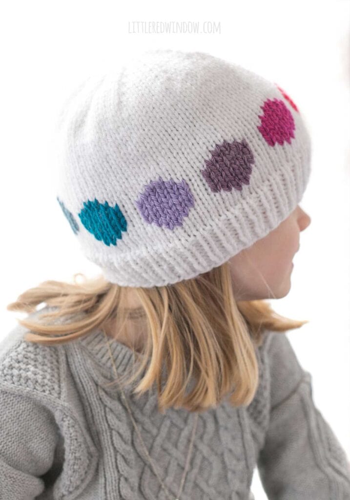 child in gray cable knit sweater wearing a white knit hat with cool rainbow colored large polka dots in a line around the middle looking off to the right
