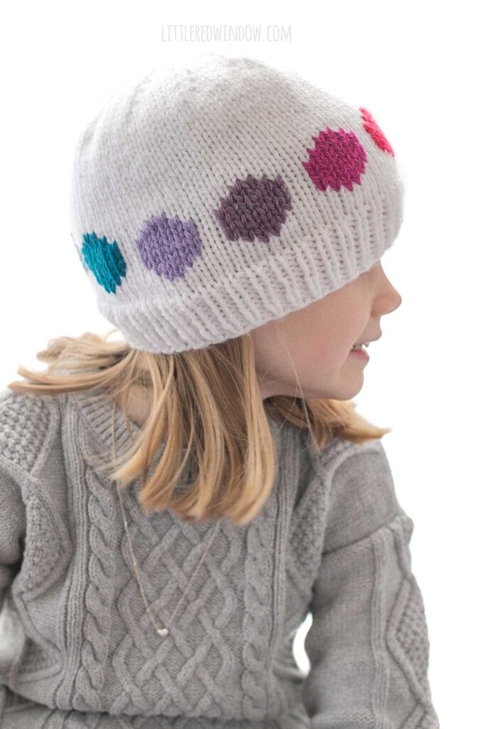child in gray cable knit sweater wearing a white knit hat with cool rainbow colored large polka dots in a line around the middle looking off to the right