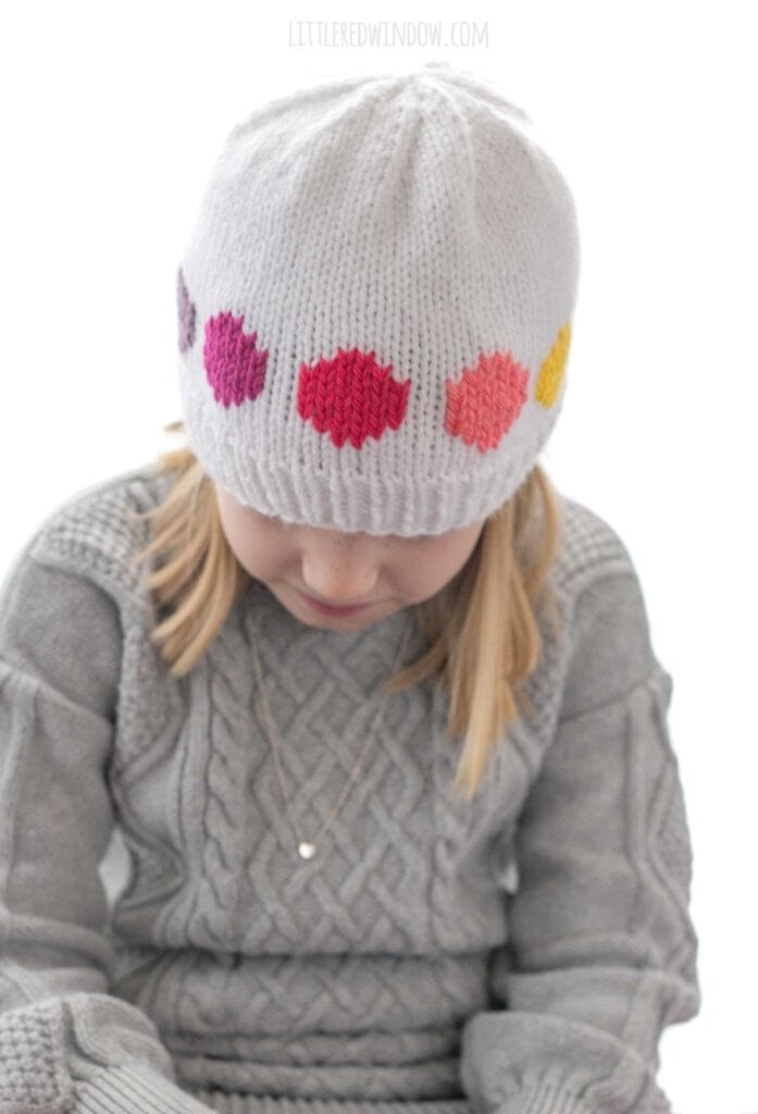 front view of girl wearing white knit rainbow polka dot hat knitting pattern wearing a gray sweater and looking down at their lap
