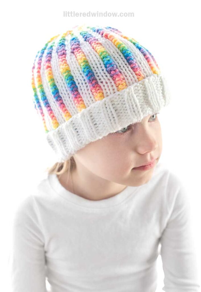 A child, wearing a vibrant rainbow rib hat with colorful stripes and a white ribbed brim, gazes to the side. Dressed in a crisp white shirt, the child is set against a bright, soft-focus backdrop.
