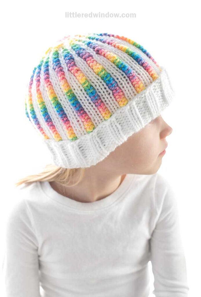 A child wearing a rainbow rib hat with colorful stripes gazes to the side. The child is dressed in a white shirt, and the background is plain white, giving a clean and bright appearance.